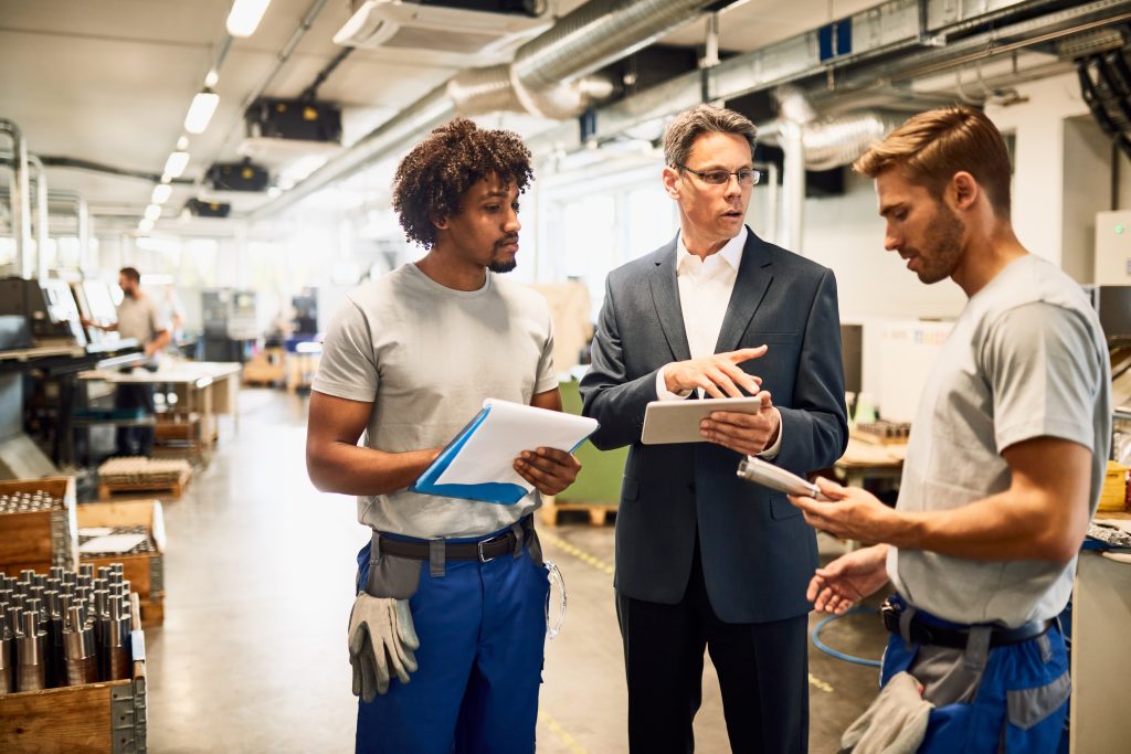 A manager in a suit reviewing a digital tablet with factory workers on the floor to address production data delays in manufacturing.