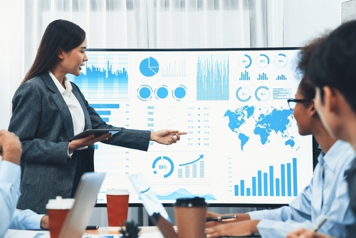 A business executive pointing at a global logistics data dashboard during a meeting focused on analyzing supply chain procurement leaks.
