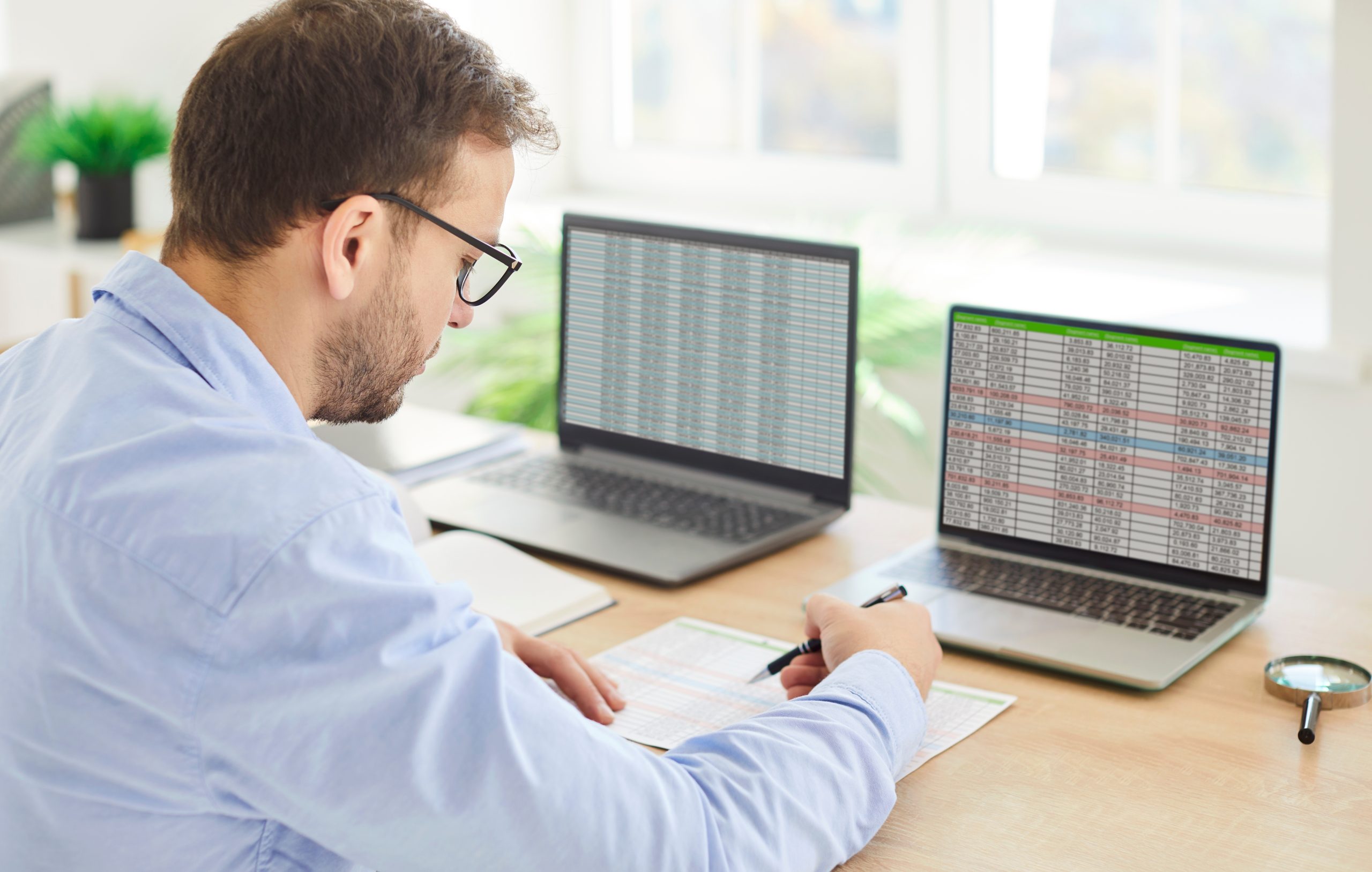 A data analyst working at a desk with two laptops and a spreadsheet, comparing regional stock levels to optimize multi-location inventory control.