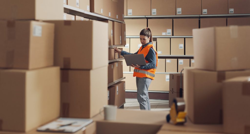 A female logistics specialist holding a laptop while checking shelves for efficient reverse supply chain inventory control.