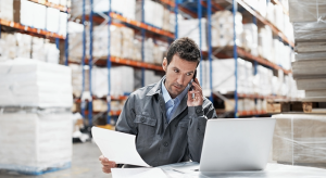 A logistics coordinator talking on the phone while looking at a laptop and documents to resolve vendor performance management gaps.